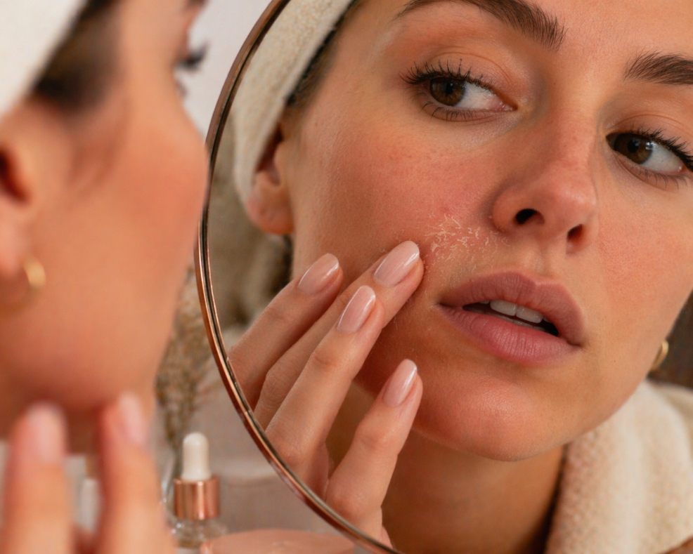 picture of woman in a bathroom mirror touching dry, flaky skin around her nose and mouth, showing signs of over-exfoliation and a damaged skin barrier