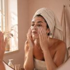picture of Woman applying exfoliating skincare in bathroom mirror with hair wrapped in towel, demonstrating a typical skincare routine that can lead to over-exfoliation