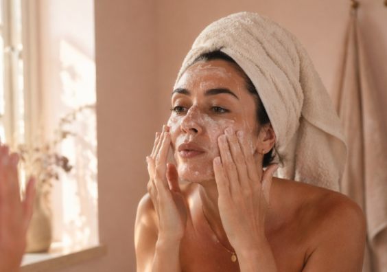 picture of Woman applying exfoliating skincare in bathroom mirror with hair wrapped in towel, demonstrating a typical skincare routine that can lead to over-exfoliation
