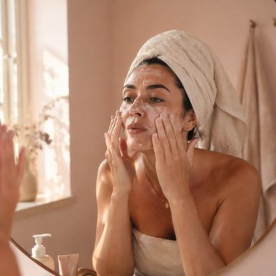 picture of Woman applying exfoliating skincare in bathroom mirror with hair wrapped in towel, demonstrating a typical skincare routine that can lead to over-exfoliation